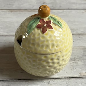 May include: A yellow ceramic lemon-shaped sugar bowl with a lid. The lid has a small orange fruit on top and green leaves and red flowers around it.