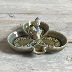 May include: A ceramic trinket dish shaped like a three-leaf clover, featuring a small figurine of a seated figure in blue and white. The dish is olive green with embossed shamrocks around the edges.