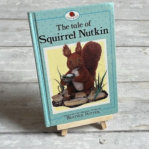 May include: A children's book titled "The tale of Squirrel Nutkin" by Beatrix Potter. The cover is light blue with white polka dots and features an illustration of a squirrel holding a nest with eggs. The Ladybird logo is at the top.