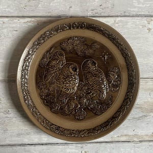 Puede incluir: Plato decorativo de cerámica marrón con tres búhos posados en una rama. El plato tiene un diseño en relieve con una estrella y el año 1970. El borde tiene un borde decorativo.