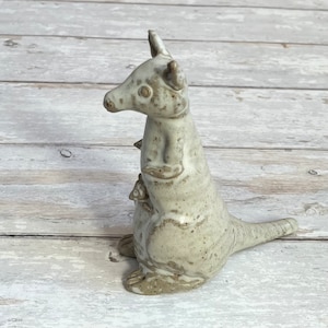 Può includere: Una statuetta in ceramica di un canguro con un piccolo nella sua tasca. La statuetta è dipinta in un colore grigio chiaro con macchie marroni.