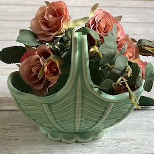 May include: A green ceramic basket with a woven design, filled with pink roses and green leaves.