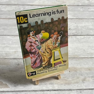 Puede incluir: Un libro infantil vintage titulado "Learning is fun" con una ilustración de dos payasos en la portada. El libro también muestra el precio "10c" y el texto "The Ladybird Key Words Reading Scheme". El libro está apoyado en un pequeño caballete de madera.