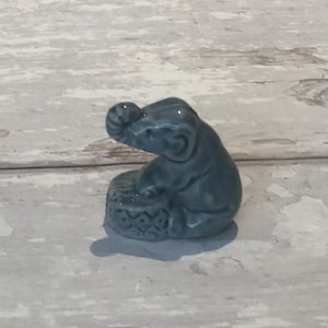 May include: A small, grey ceramic elephant figurine sitting on a base with a decorative pattern. The elephant has its trunk raised and is holding a small object in its trunk.