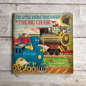 Könnte beinhalten: Ein Vintage-Kinderbuch mit dem Titel "The Little Engine That Could & The Big Chase" mit einer Illustration einer blauen Lokomotive, die einen roten Waggon mit einem Clown darauf zieht. Das Buch ist Teil der Reihe "All Aboard Books".
