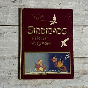 Peut inclure: Un livre vintage intitulé "Sindbad's First Voyage" avec une couverture bordeaux et des lettres dorées. La couverture présente des illustrations de deux pingouins vêtus d'habits d'hiver qui font du traîneau et une lune croissante dans le ciel.