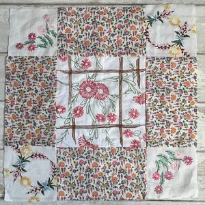 Könnte beinhalten: Ein Patchwork-Quilt-Quadrat, das aus neun kleineren Quadraten besteht. Die Quadrate zeigen florale Muster und gestickte Designs in Rosa, Gelb und Blau auf weißem Hintergrund. Einige Quadrate haben Blumenkränze, andere gestickte Blumen.