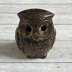Op de afbeelding: Een keramische spaarpot in de vorm van een uil, in bruin- en beigetinten. De uil heeft grote, ronde oogkassen en een gleuf aan de bovenkant om munten in te werpen. Het oppervlak heeft een getextureerd, gespikkeld uiterlijk.