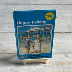 May include: A blue book with the title "Happy holiday" and the number "7a" in a yellow circle. The book cover features a colorful illustration of a pier, beach, and children playing. The book is part of the "Key Words Reading Scheme".