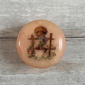 May include: A round, pink, alabaster trinket box with a painted image of a girl in a brown hat sitting on a fence. The girl is wearing a purple shirt and green pants. The background is a light pink with green leaves and flowers.