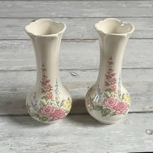 Pode incluir: Dois vasos de cerâmica creme com um design floral com rosas cor-de-rosa, flores azuis e amarelas e folhas verdes. Os vasos têm uma borda ondulada e uma base bulbosa.