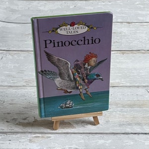 Könnte beinhalten: Ein lilafarbenes Buch mit grünem Rücken, betitelt "Well-Loved Tales Pinocchio", mit einer Illustration von Pinocchio, der auf einem Tauben über einem blauen Meer mit einem Segelboot in der Ferne reitet.