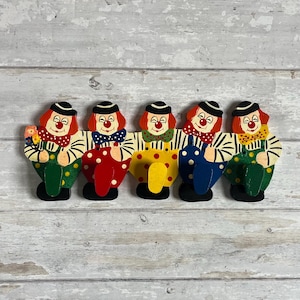 Puede incluir: Una fila de cinco figuras de payasos de madera con detalles pintados. Cada payaso tiene una peluca roja, un sombrero negro y una cara pintada. Los payasos visten trajes de varios colores, incluyendo lunares y rayas, y sostienen accesorios.