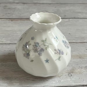 May include: A white ceramic vase with a scalloped rim and a floral pattern of purple and blue flowers. The vase has a bulbous shape and is ideal for displaying a small bouquet of flowers.