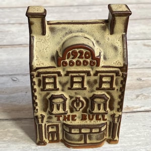 May include: A ceramic figurine of a building with a brown and tan color scheme. The building has a sign that reads "THE BULL" and the number "1920" above the windows.