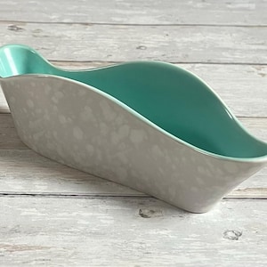 Op de afbeelding: Een keramische serveerschaal met een lichtgrijze buitenkant en een turquoise binnenkant. De schaal heeft een gebogen, bootachtige vorm met een opstaande rand aan één uiteinde. De buitenkant heeft een gespikkeld patroon.