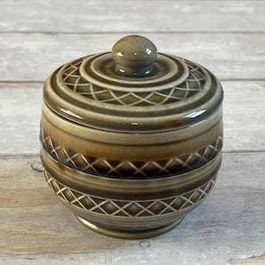 Può includere: Un piccolo barattolo rotondo in ceramica con coperchio. Il barattolo è principalmente marrone con un motivo decorativo di linee che si intersecano. Il coperchio ha un piccolo pomello arrotondato. Il barattolo poggia su una piccola base circolare.