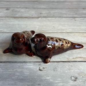 May include: Two ceramic seal figurines. The seals are brown with a glossy finish and a speckled pattern on their backs. They are lying down, with their heads raised. The background is a light gray wooden surface.