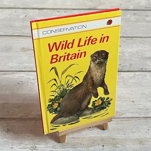 Könnte beinhalten: Ein Buch mit dem Titel "Wild Life in Britain" mit gelbem Einband und roten Akzenten. Das Cover zeigt einen braunen Otter und grüne Pflanzen. Das Buch steht auf einer kleinen Holzstaffelei.
