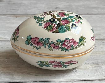 Könnte beinhalten: Eiförmige Schmuckschatulle in Creme mit floralem Design. Der Deckel zeigt einen Baum mit rosa Blüten und grünen Blättern. Der Sockel hat eine goldene Zierleiste und ein passendes Blumenmuster.