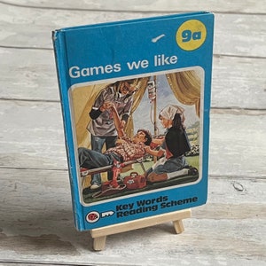 May include: A blue book with the title "Games we like" and a red ladybug logo. The book is on a small wooden easel. The cover features an illustration of children playing a game.