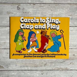 May include: A yellow book titled "Carols to Sing, Clap and Play" with illustrations of children in Christmas costumes playing musical instruments. The book is a companion to the descant recorder tuition books by Heather Cox and Garth Rickard.