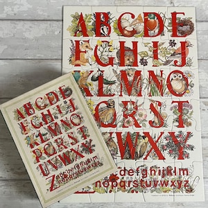 May include: A colorful alphabet puzzle with uppercase letters in red, each letter adorned with illustrations of animals and plants. A matching box with the alphabet is next to the puzzle. The puzzle is on a light wood-grain surface.