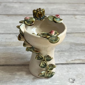 Może przedstawiać: Ceramiczny wazon z motywem żaby z kremową podstawą. Wazon posiada żabę na górze, liście lilii wodnych i liście. Liście lilii wodnych mają różowe akcenty. Liście i żaba są zielone i brązowe.