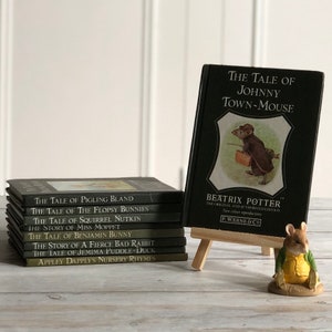 May include: A stack of ten black hardback books with gold lettering on the spines. The titles of the books are "The Tale of Pigling Bland", "The Tale of the Flopsy Bunnies", "The Tale of Squirrel Nutkin", "The Story of Miss Moppet", "The Tale of Benjamin Bunny", "The Story of a Fierce Bad Rabbit", "The Tale of Jemima Puddle-Duck", and "Appley Dappley's Nursery Rhymes". A black hardback book with a gold title, "The Tale of Johnny Town-Mouse", is standing upright on a wooden easel. A small figurine of a mouse wearing a green jacket and yellow shirt is standing on a wooden surface.