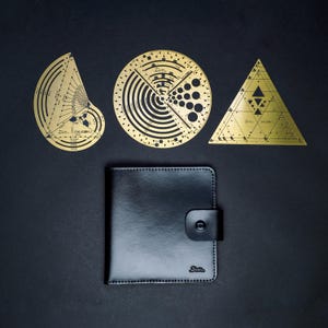 May include: A black leather wallet with a snap closure and three brass geometric tools. The tools include a semi-circle, a circle, and a triangle, all with intricate designs and text. The tools are made of brass.