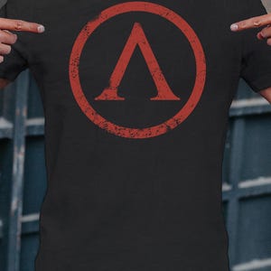 Puede incluir: Camiseta negra con un símbolo espartano rojo desgastado, una letra lambda dentro de un círculo. El gráfico tiene un aspecto desgastado. La camiseta está hecha de tela oscura y se usa con vaqueros azules.