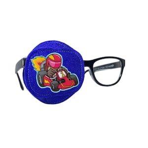 Peut inclure: Patch oculaire bleu avec une image de dessin animé d'un pilote de course portant un casque et conduisant une voiture de course rouge avec une flamme à l'arrière. Le patch oculaire est attaché à une paire de lunettes à monture noire.