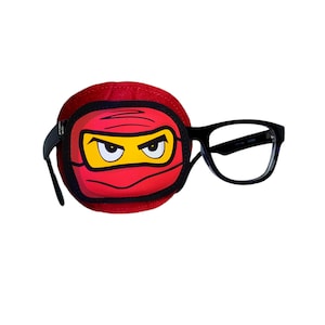 Op de afbeelding: Een rood ooglapje met een cartoon ninja-gezicht erop. Het ooglapje is bevestigd aan een zwarte bril.