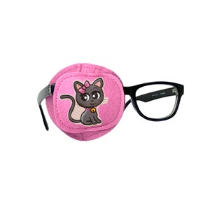 Könnte beinhalten: Eine rosa Augenklappe mit einem Cartoon-Katzen-Design, befestigt an einer schwarzen Brille. Die Katze hat eine rosa Schleife, eine goldene Glocke und einen grauen Körper. Die Brille hat schwarze Rahmen.