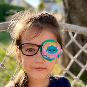 Könnte beinhalten: Ein junges Mädchen mit Brille und einem bunten Augenpflaster mit einer Comicfigur darauf. Das Augenpflaster ist grün, rosa und blau mit einer flauschigen weißen Figur mit einer rosa Nase und blauen Augen. Das Mädchen lächelt und schaut in die Kamera.