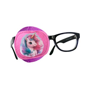 Könnte beinhalten: Ein rosa Augenpflaster mit einem Einhorn-Design. Das Augenpflaster ist an einer schwarzen Brille befestigt.