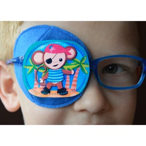 Puede incluir: Un parche para el ojo azul con un diseño de mono pirata. El mono lleva un sombrero rojo con una calavera y huesos cruzados, una camisa a rayas azul y blanca y un parche para el ojo negro. El mono está de pie en una playa con palmeras.
