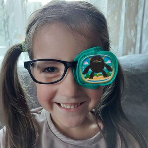 Könnte beinhalten: Ein junges Mädchen mit Brille und einem grünen Augenpflaster mit einem Cartoon-Monster-Design. Das Pflaster bedeckt ihr rechtes Auge.