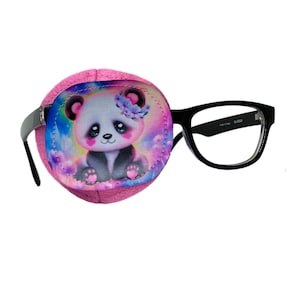 Op de afbeelding: Roze oogpleister met een kleurrijk pandabeerontwerp. De oogpleister is bevestigd aan een bril met een zwart montuur.