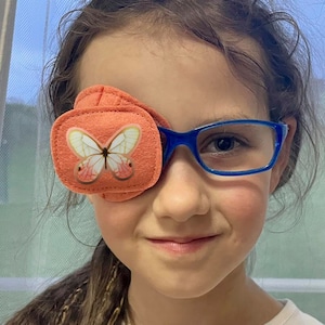 Könnte beinhalten: Ein junges Mädchen trägt eine blaue Brille mit einem orangefarbenen Augenpflaster, das mit einem Schmetterlingsmotiv verziert ist.
