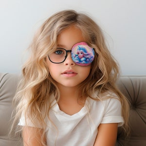 Puede incluir: Una niña joven con cabello largo y rubio lleva gafas con montura negra y un parche para el ojo rosa con un diseño de unicornio. El parche para el ojo tiene una imagen de un unicornio con una corona y un arcoíris.