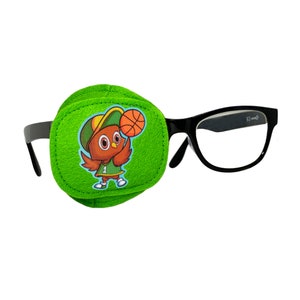 Op de afbeelding: Een groene ooglap met een cartoon uil die een baseballpet draagt en een basketbal vasthoudt. De ooglap is bevestigd aan een zwarte bril.