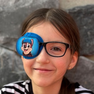Op de afbeelding: Een jong persoon draagt een bril en een blauw ooglapje met een cartoonhondontwerp. Het ooglapje bedekt het linkeroog.