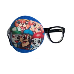 Può includere: Cerotto per l'occhio blu con un'immagine di cartone animato colorata di cinque cuccioli della serie di cartoni animati Paw Patrol. Il cerotto per l'occhio è attaccato a un paio di occhiali con montatura nera.