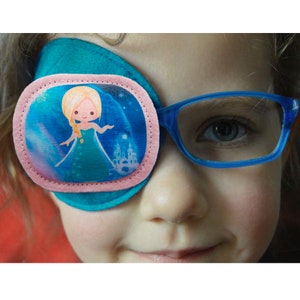 Puede incluir: Un parche para el ojo turquesa con un borde rosa y una imagen impresa de una princesa de dibujos animados con un vestido azul. La princesa está de pie frente a un castillo azul con estrellas en el fondo.