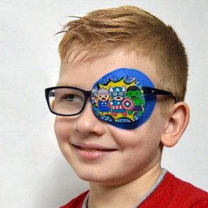 Puede incluir: Un parche para el ojo azul con una imagen de dibujos animados de superhéroes. El parche lo lleva puesto un joven con el pelo rubio y gafas.