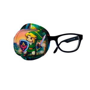 Puede incluir: Un parche en el ojo con una colorida ilustración de un personaje de dibujos animados con una espada y un escudo. El parche en el ojo está sujeto a un par de gafas con montura negra. El personaje viste de verde y empuña una espada y un escudo.