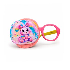 Puede incluir: Un parche ocular de tela rosa para gafas con una ilustración colorida de un conejo de dibujos animados con orejas de arcoíris que sostiene un corazón rosa. El parche está unido a una montura de gafas de plástico rosa con patillas amarillas.