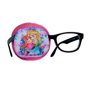 Może przedstawiać: Różowy łatkę na oko z nadrukowanym obrazkiem dwóch kreskówkowych księżniczek, które się tulą. Łatka na oko jest przymocowana do czarnych okularów.