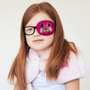 Può includere: Un cerotto per l'occhio rosa con un design di Minnie Mouse. Il cerotto viene indossato da una bambina con i capelli lunghi e rossi.